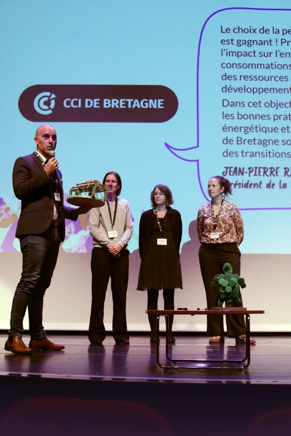 Un homme tient un micro et un trophée artisanal sur scène aux côtés de trois femmes, devant un écran affichant le logo de la CCI de Bretagne.