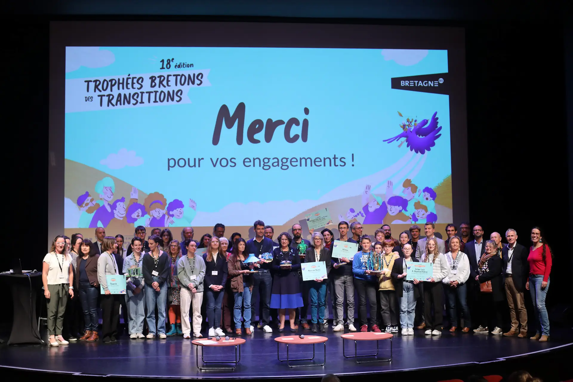Grand groupe de lauréats et organisateurs réunis sur scène devant un écran projetant la mention « Merci pour vos engagements » lors de la 18e édition des Trophées bretons des transitions.