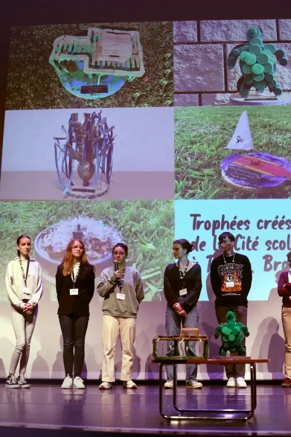 Groupe d’élèves debout sur scène devant des images projetées des trophées qu’ils ont conçus dans le cadre des Trophées bretons des transitions.
