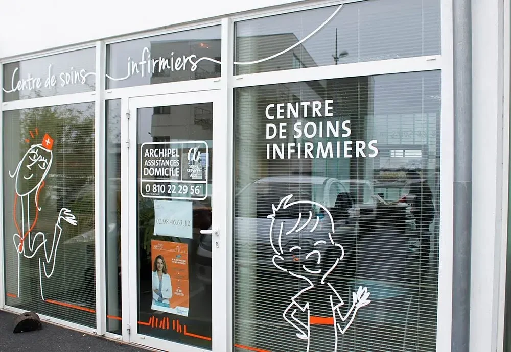 Vue de la façade vitrée d’un centre de soins infirmiers Archipel Santé, ornée de dessins illustrant un soignant et un enfant, avec les informations de contact affichées sur la porte.