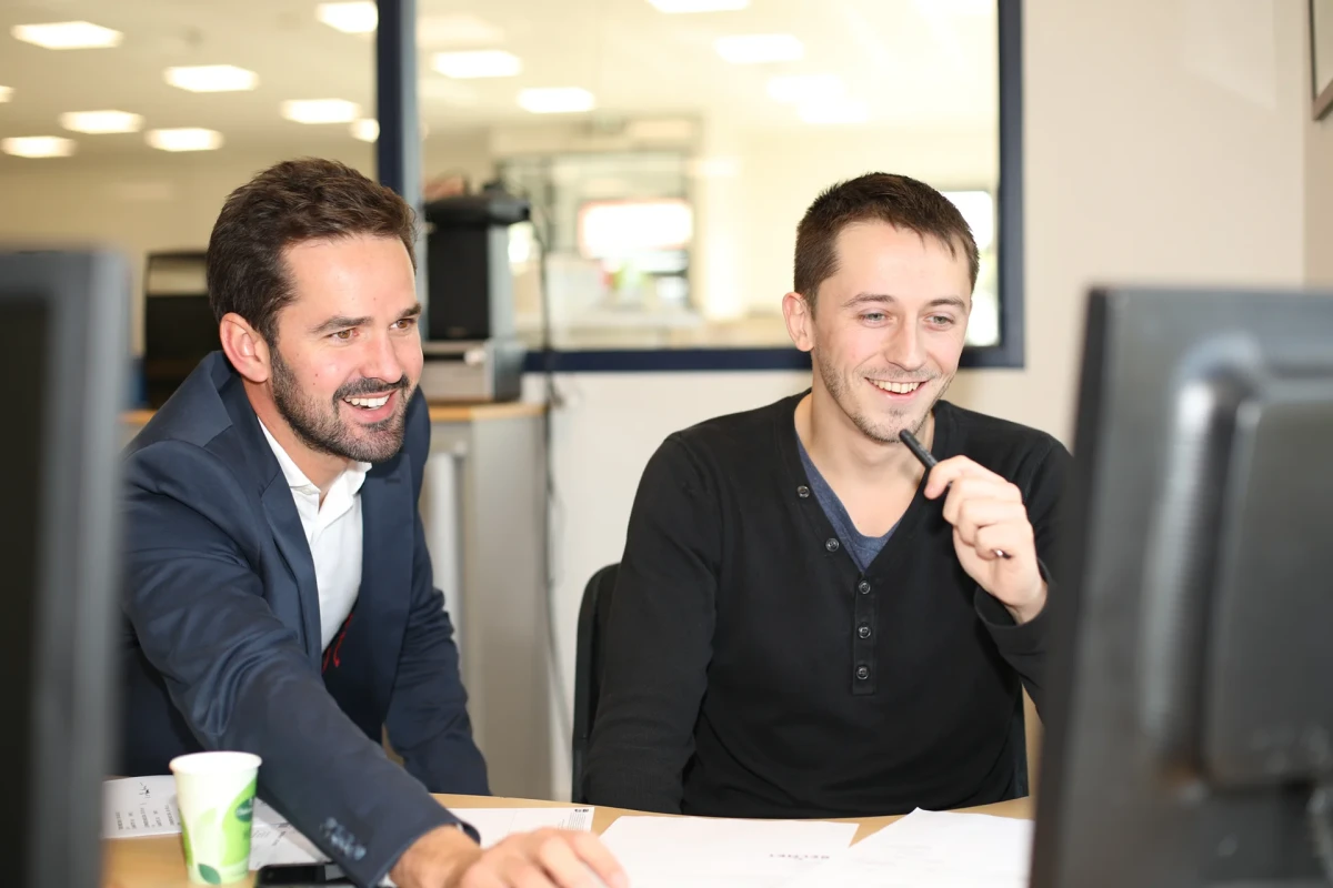 Deux collaborateurs de Belmet souriants travaillent ensemble devant un écran d’ordinateur dans un bureau lumineux, illustrant la collaboration et la cohésion interne de l’entreprise.