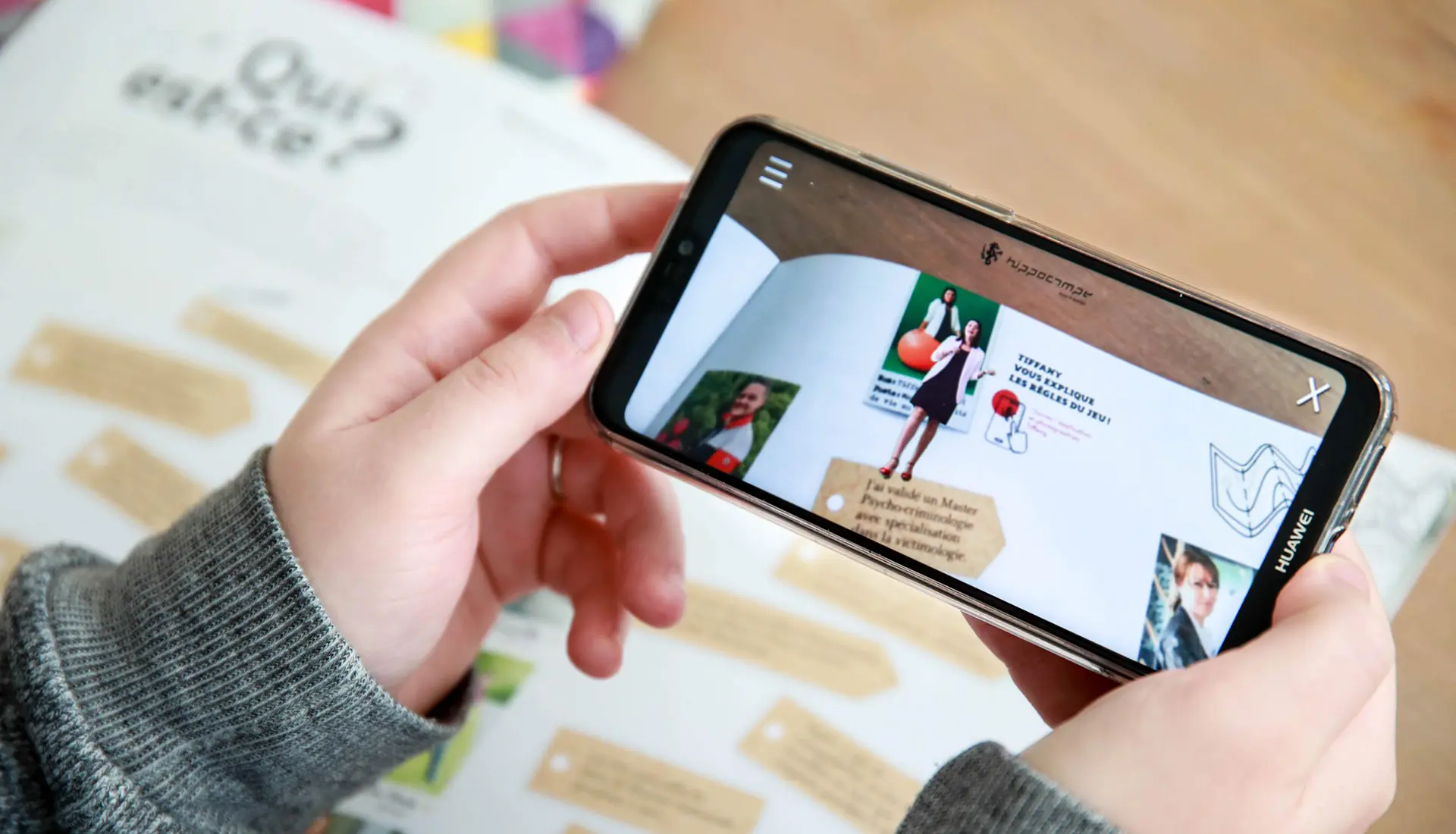 Main tenant un smartphone affichant une animation en réalité augmentée intégrée à un livret de présentation, illustrant une expérience interactive développée pour les collaborateurs de Cadiou.