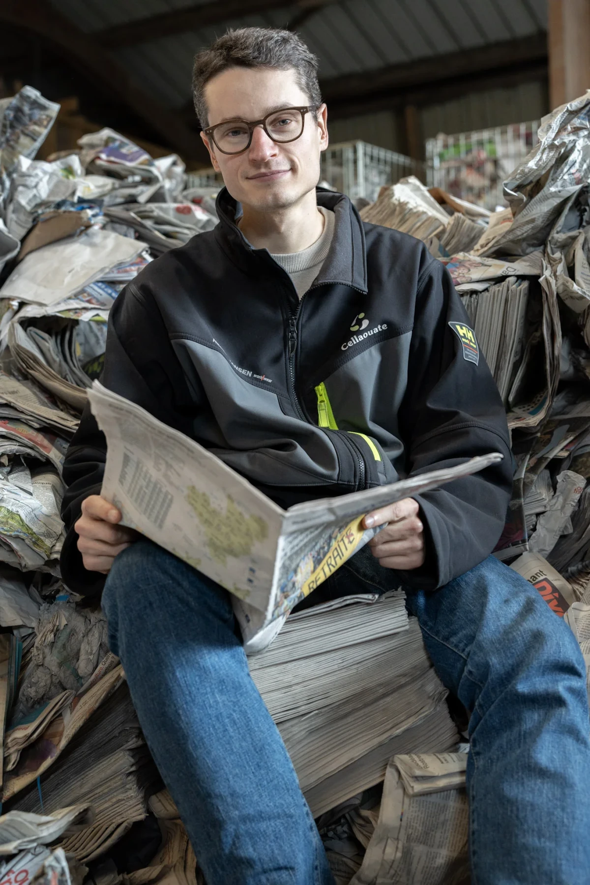 Portrait d’un collaborateur de Cellaouate assis sur une pile de journaux recyclés, lisant un quotidien, symbole de la maîtrise du processus de transformation du papier en matériau isolant écologique.