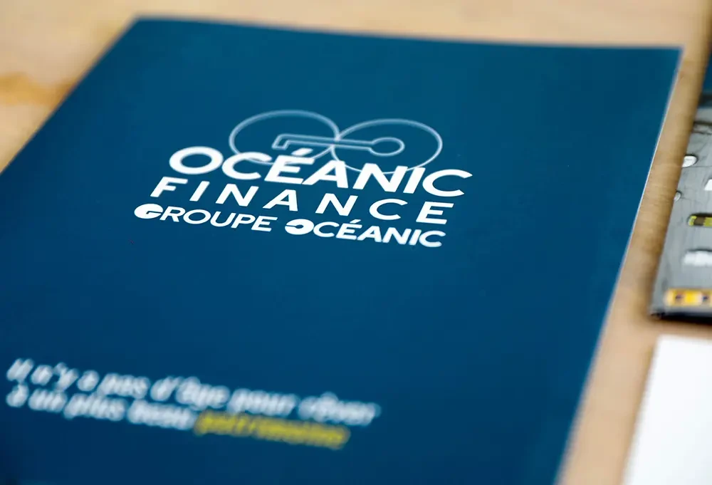 Gros plan sur la couverture bleu marine de la plaquette Océanic Finance, affichant le logo blanc et le slogan « Il n’y a pas d’âge pour rêver à un plus beau patrimoine ».