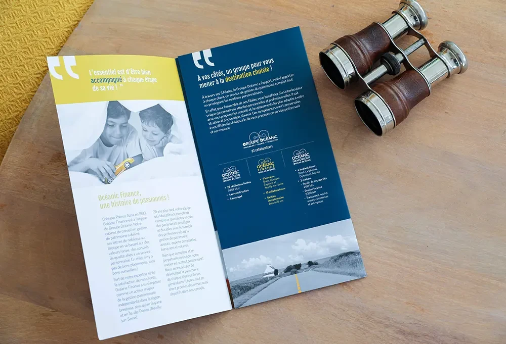 Vue de la plaquette Océanic Finance ouverte, posée sur une table en bois. À gauche, une photo en noir et blanc de deux enfants sous une couverture. À droite, du texte blanc sur fond bleu marine décrivant les services et la mission du groupe. Jumelles anciennes posées à côté.