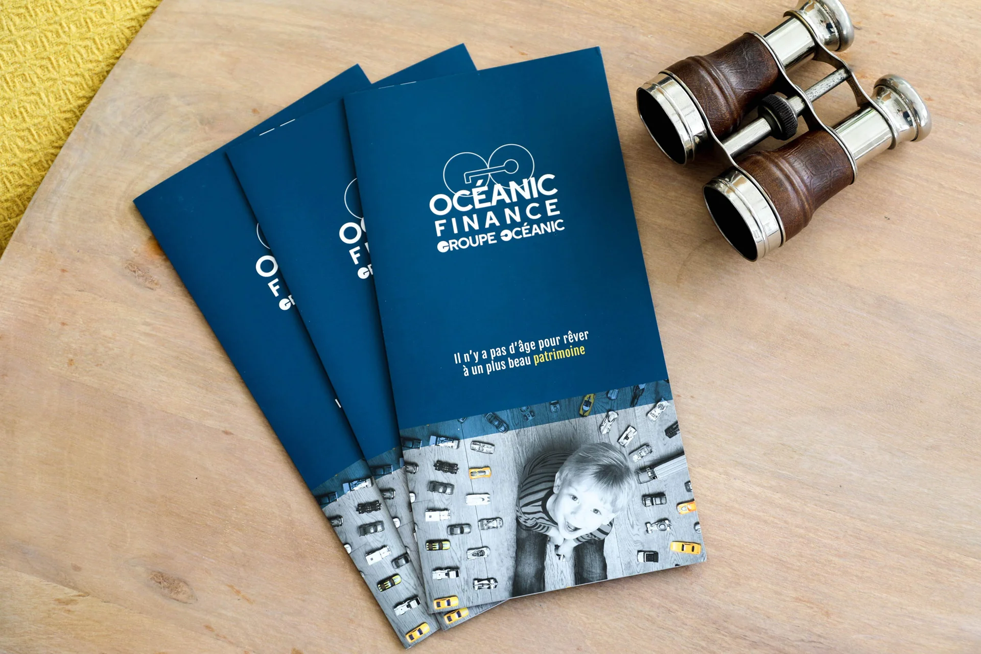 Trois plaquettes institutionnelles bleu marine d’Océanic Finance disposées en éventail sur une table en bois clair, accompagnées de jumelles anciennes. Le visuel de couverture présente le logo de l’entreprise et une photo en noir et blanc d’un enfant entouré de petites voitures.