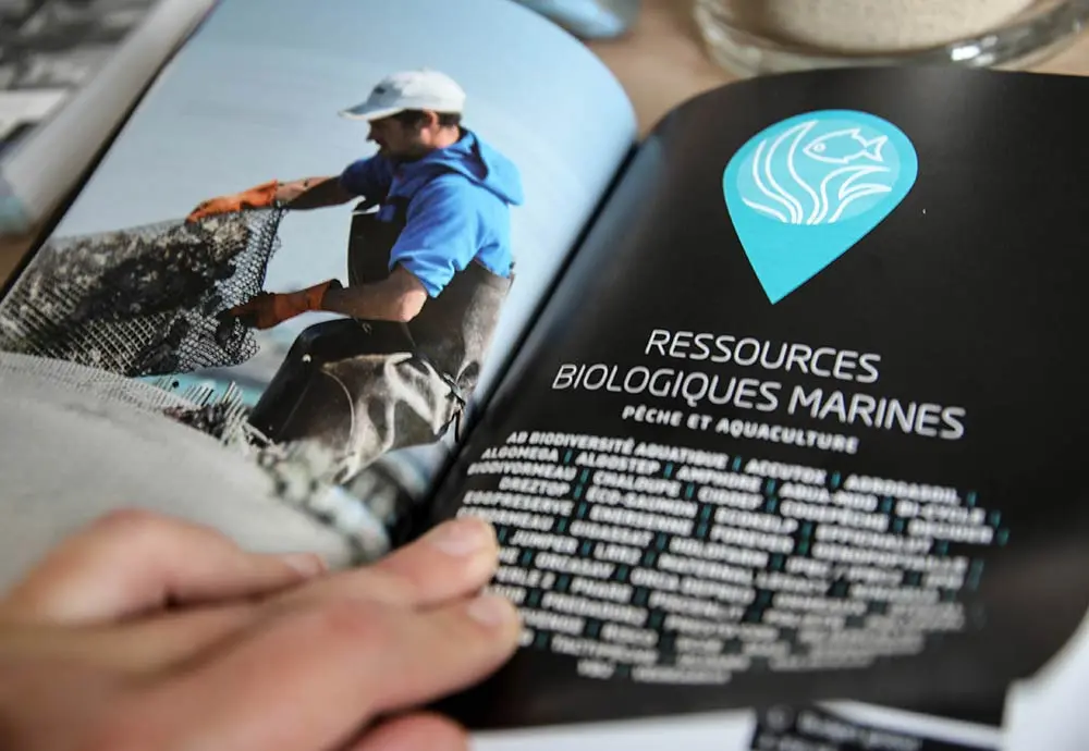 Vue intérieure du Livre de Bord du Pôle Mer présentant une section sur les ressources biologiques marines. À gauche, une photo d’un ostréiculteur au travail, à droite, une page noire avec pictogramme bleu et titre blanc.