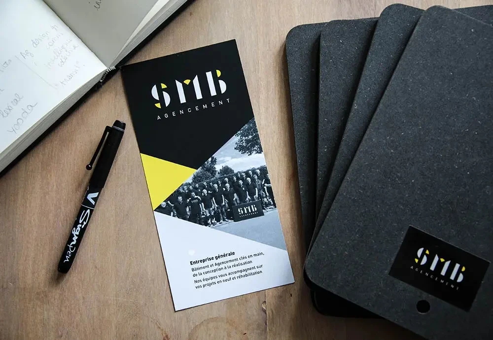 Dépliant SMB Agencement au graphisme noir, blanc et jaune avec photo d’équipe, posé sur un bureau