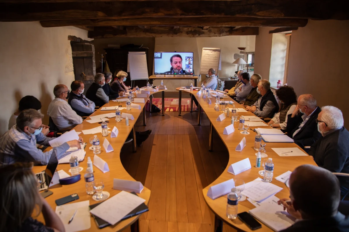 Réunion d’un groupe de personnes assises autour de deux grandes tables en bois formant un U, avec une visioconférence projetée sur un écran au fond de la salle.