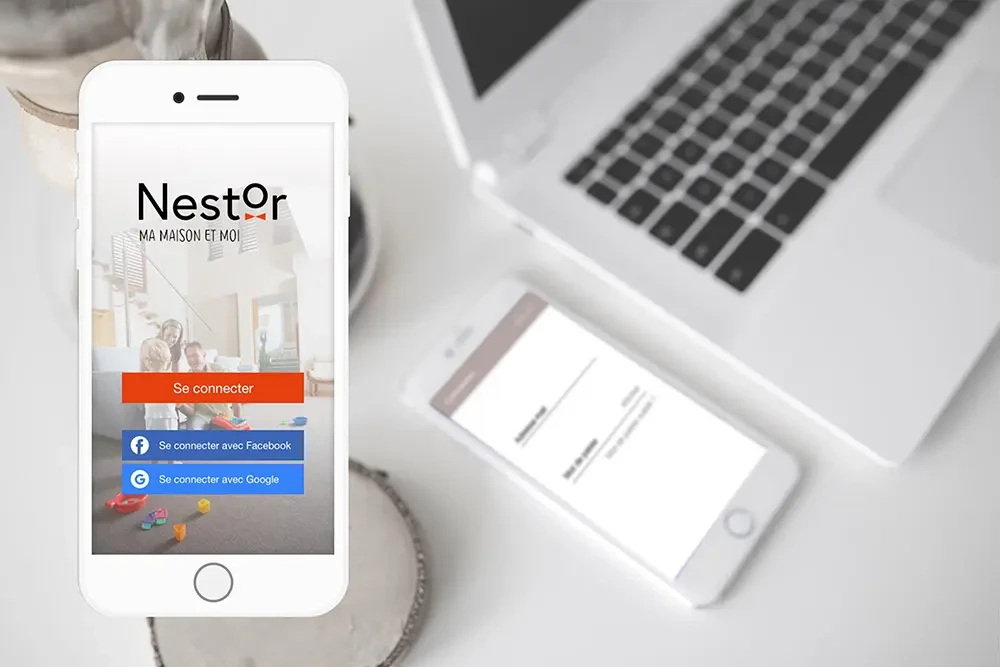 Écran d’accueil de l’application mobile Nestor affiché sur smartphone, avec options de connexion Facebook et Google, devant un ordinateur portable