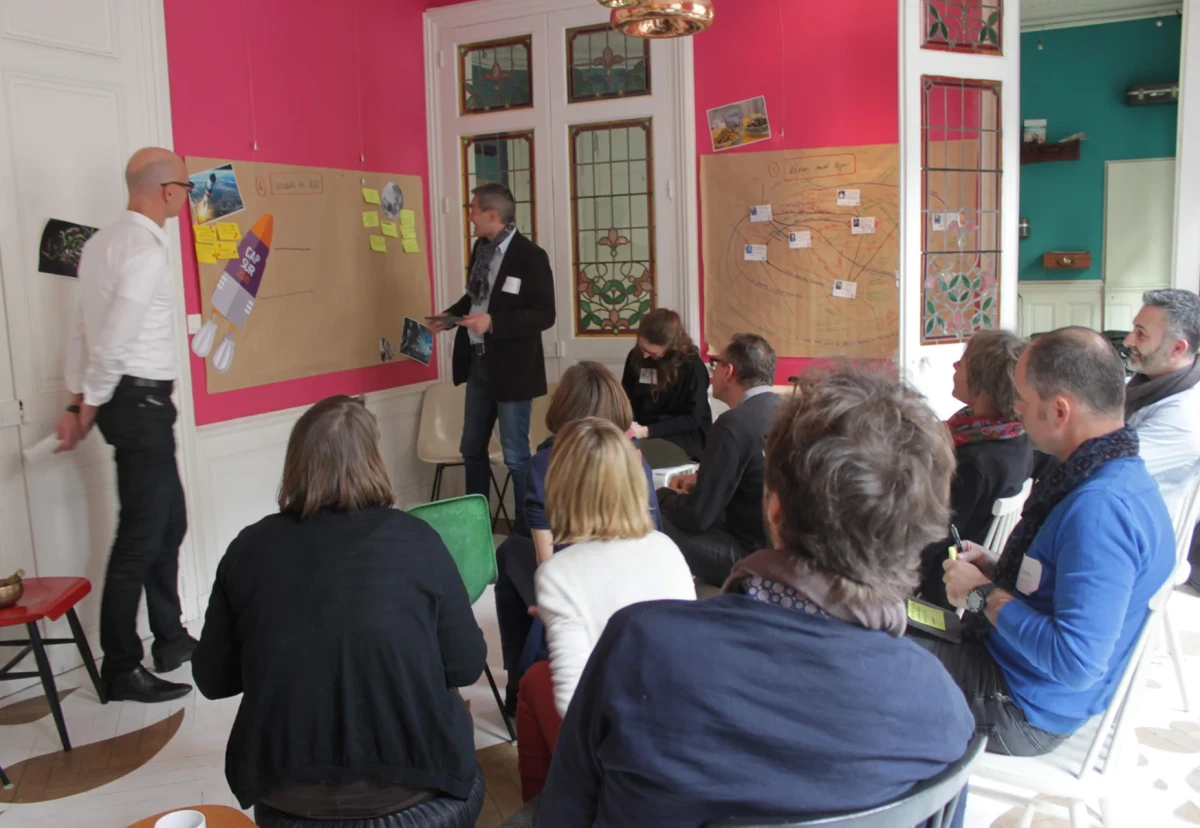 Un groupe de personnes assises participe à un atelier d’intelligence collective dans une salle colorée à Rennes, avec deux animateurs présentant des cartes et des post-it sur de grands panneaux kraft accrochés au mur.