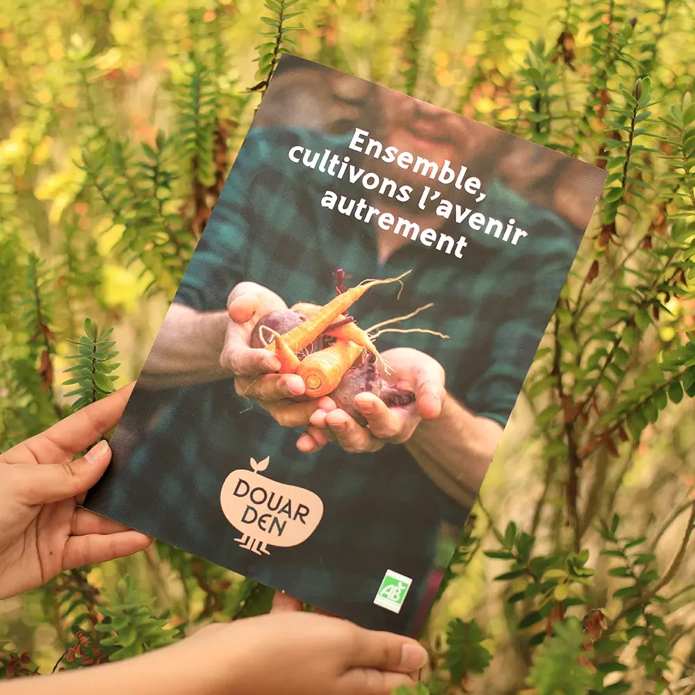 Couverture du dépliant Douar Den tenu en main, illustrant un maraîcher tenant des légumes bio colorés, avec le slogan « Ensemble, cultivons l’avenir autrement », dans un décor naturel de feuillage.