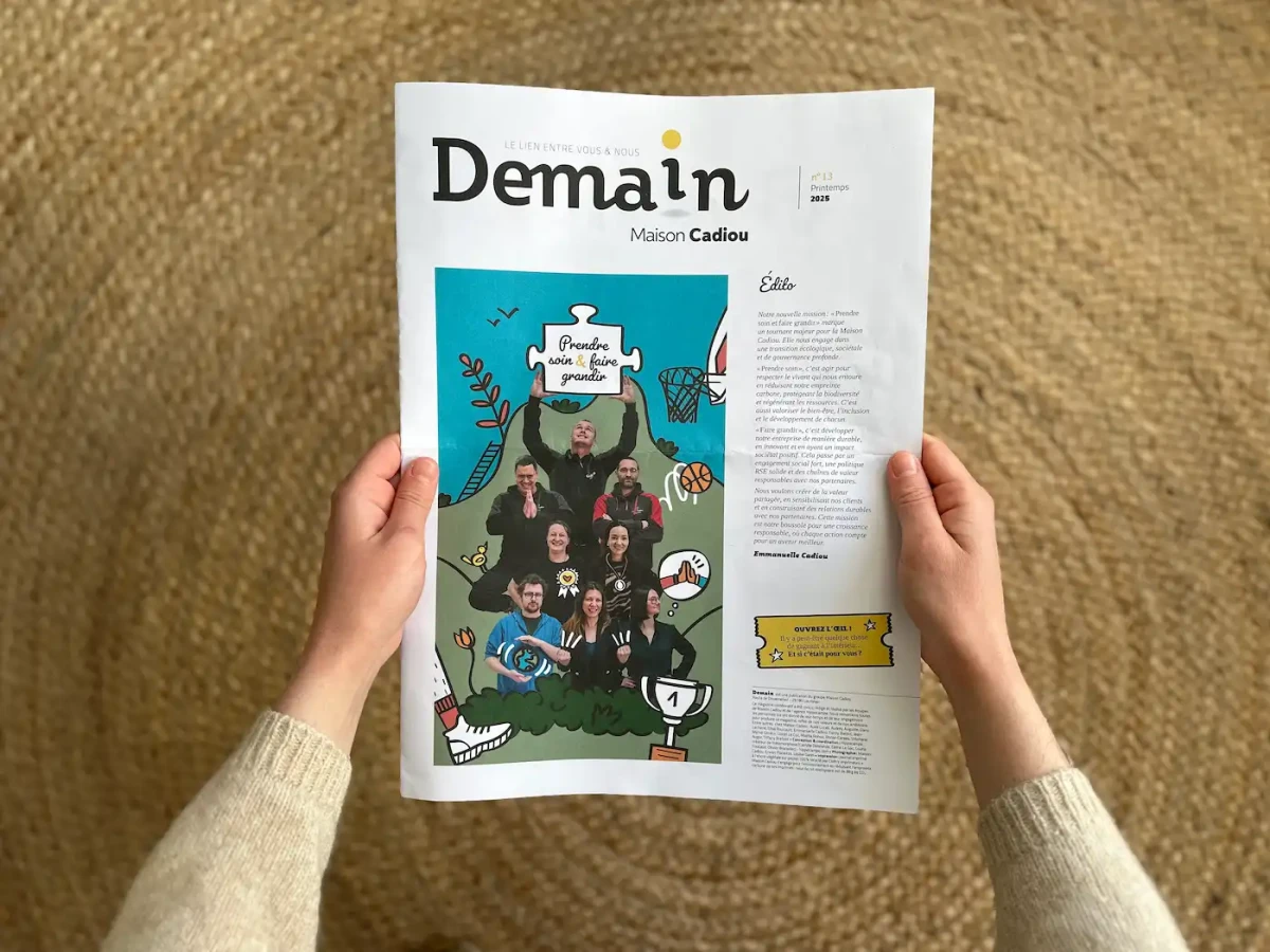 Une personne tient entre ses mains le journal interne « Demain – Maison Cadiou », dont la une colorée met en scène des collaborateurs illustrés dans un univers graphique joyeux, autour du thème « Prendre soin & faire grandir ».