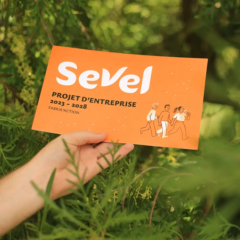 Couverture orange du livret « Sevel projet d’entreprise 2023-2028 » tenue en main, dans un cadre végétal, illustrant une démarche collective et humaine portée par l’entreprise.